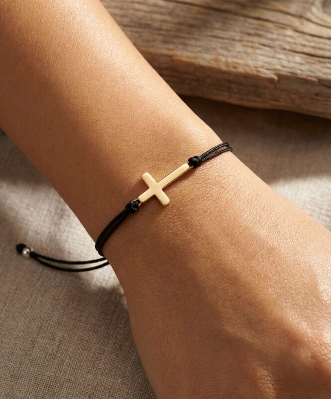 Pormenor de uma mulher a usar no pulso uma pulseira de fio preto com uma cruz dourada deitada.